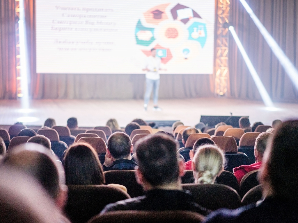 EVH Events' successful event and event production team recommend a practice run of the event presenter on stage speaking in front of a large presentation screen and stage lighting, viewed from the perspective of an audience seated in an auditorium.