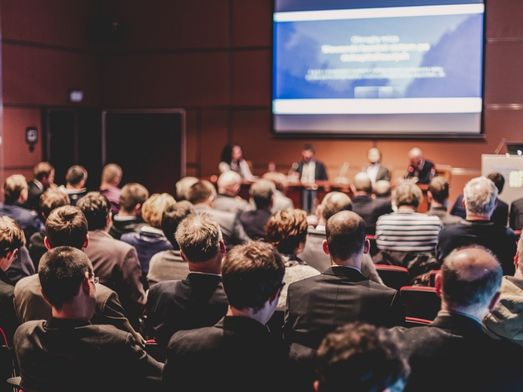 An elevated, rear-perspective view looking over a large audience of people seated in a modern auditorium during a professional conference presentation, focusing on a panel and screen at the front.